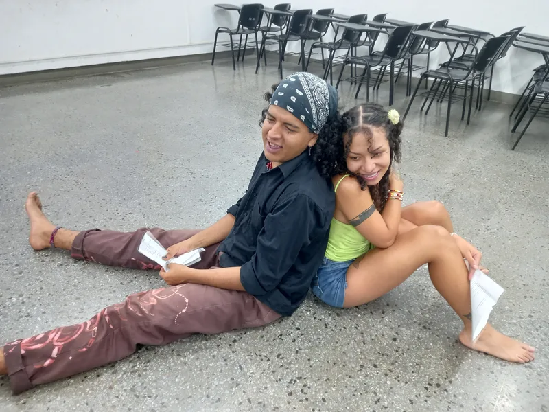 Dos integrantes sentados espalda con espalda en el piso ríen con sus textos en la mano