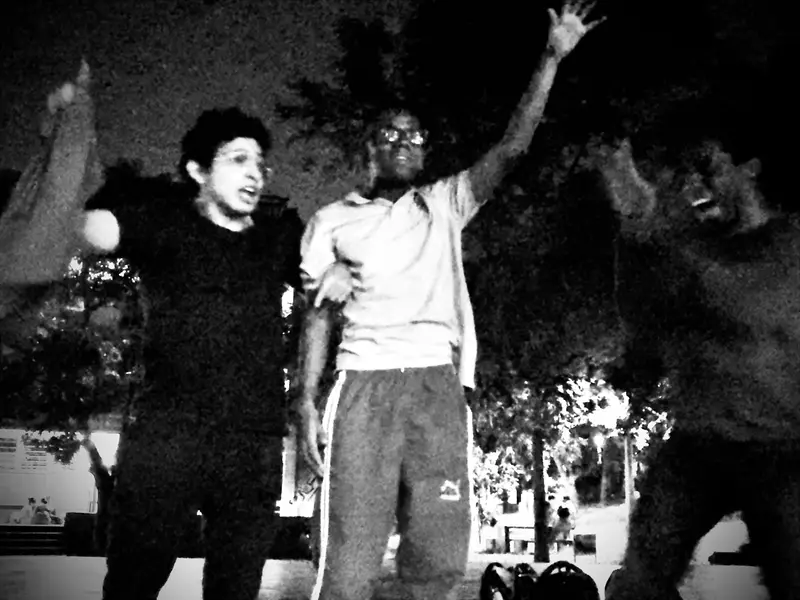En blanco y negro, un integrante levanta el brazo con alegría junto a sus compañeros en la plaza, de noche